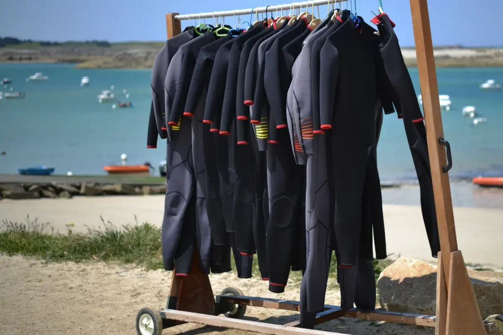 Wetsuits on a rack near the water with a mix of zipperless and zippered wetsuits